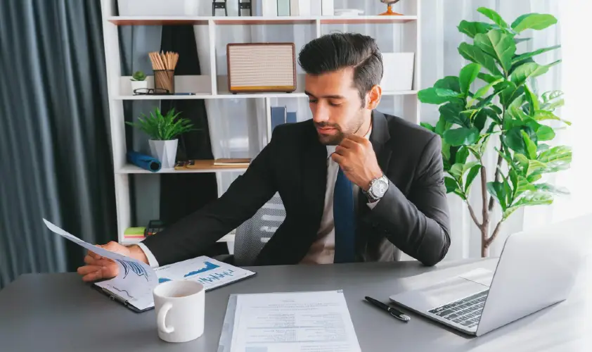 A man in a suit sits at a desk with a cup of coffee and a laptop.