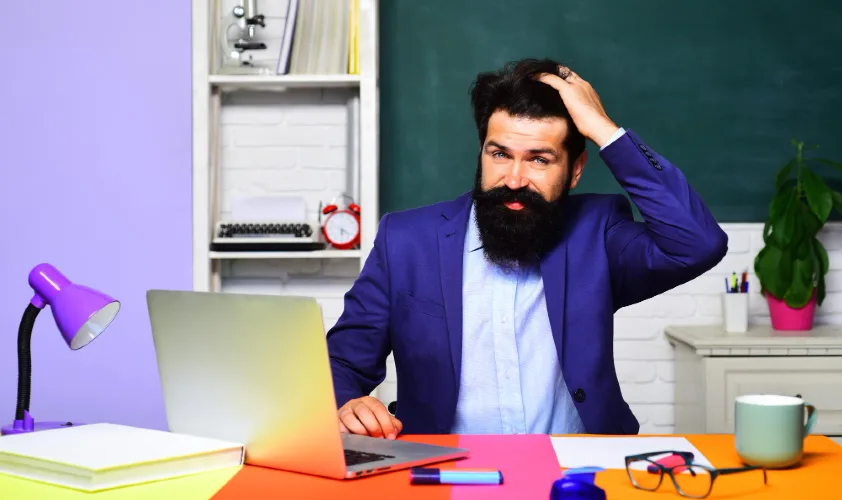 A man in suit sitting on workdesk and one hand on his head while other hand is on laptap
