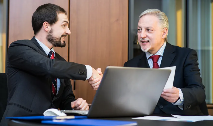 Business people handshake with papers in hand and laptop on table